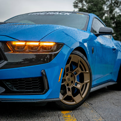 Close-up of a blue Ford Mustang Dark Horse featuring EKW GTD-1B forged aluminum wheels in a bronze finish. The aggressive multi-spoke design of the wheel pairs with a high-performance Brembo brake caliper in blue. The Mustang’s signature LED headlights are illuminated, casting an amber glow, while water droplets on the car's surface enhance its striking appearance. The front bumper and black aerodynamic accents further emphasize the vehicle’s track-ready design.