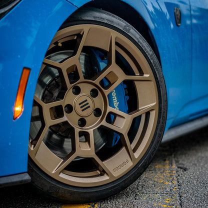 Close-up of the front wheel on a blue Ford Mustang Dark Horse, featuring EKW GTD-1B forged aluminum wheels in a bronze finish. The multi-spoke design provides strength and lightweight performance, while the engraved 'FORGED' marking highlights its premium construction. Behind the wheel, a high-performance Brembo brake caliper in blue contrasts sharply with the bronze finish. The wet pavement and water droplets on the car add a dynamic touch to the scene, emphasizing its aggressive and track-ready stance.