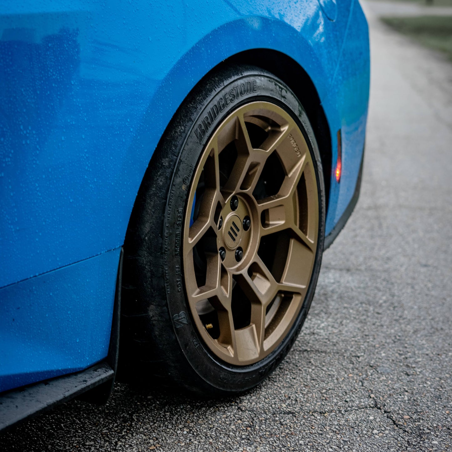 Close-up of the rear wheel on a blue Ford Mustang, featuring EKW GTD-1B forged aluminum wheels in a bronze finish. The wheel's bold multi-spoke design enhances its aggressive stance, paired with a high-performance Bridgestone tire. The Mustang's body is covered in water droplets, highlighting the car's sleek paintwork. The black side skirt and illuminated side marker add to the sporty aesthetic, with the textured pavement providing a contrasting backdrop.
